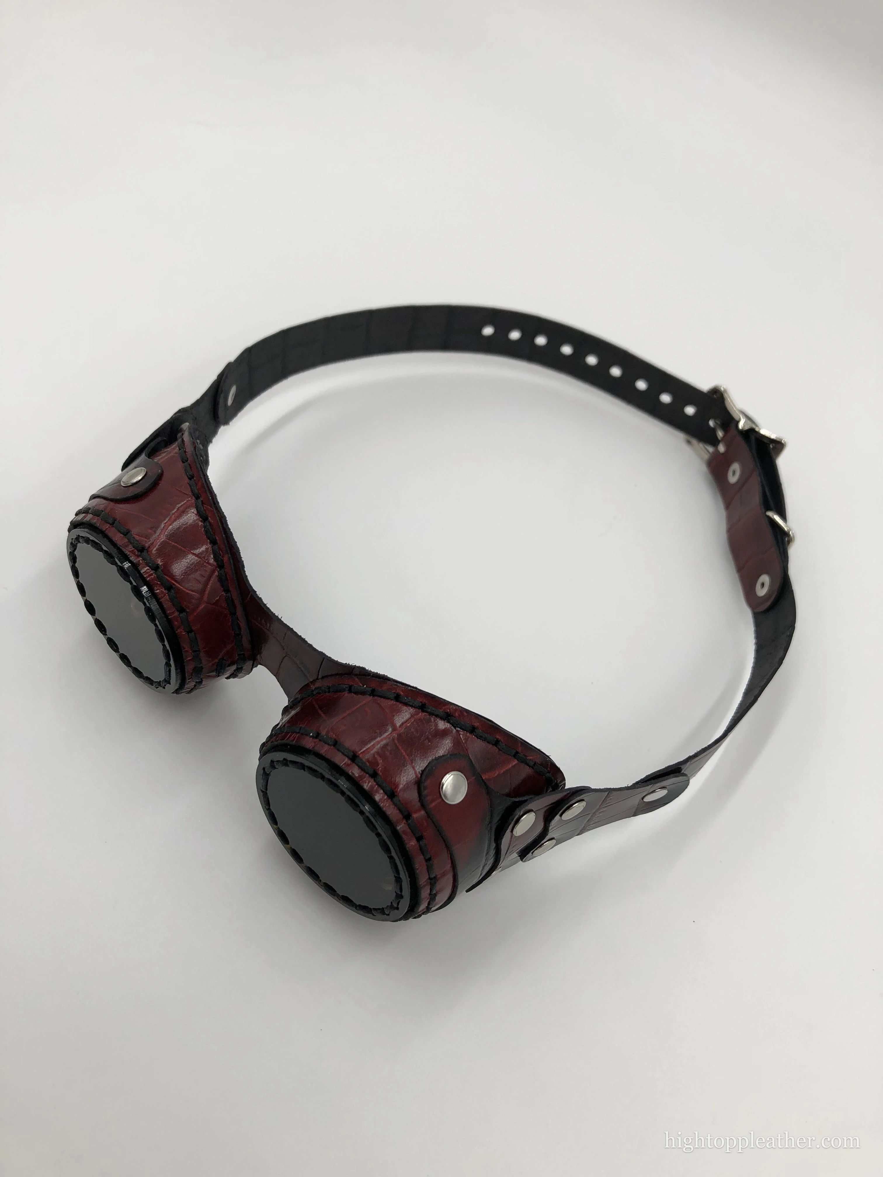 Oxblood leather aviator goggles with embossed croc texture, contrast stitching, and an adjustable strap.