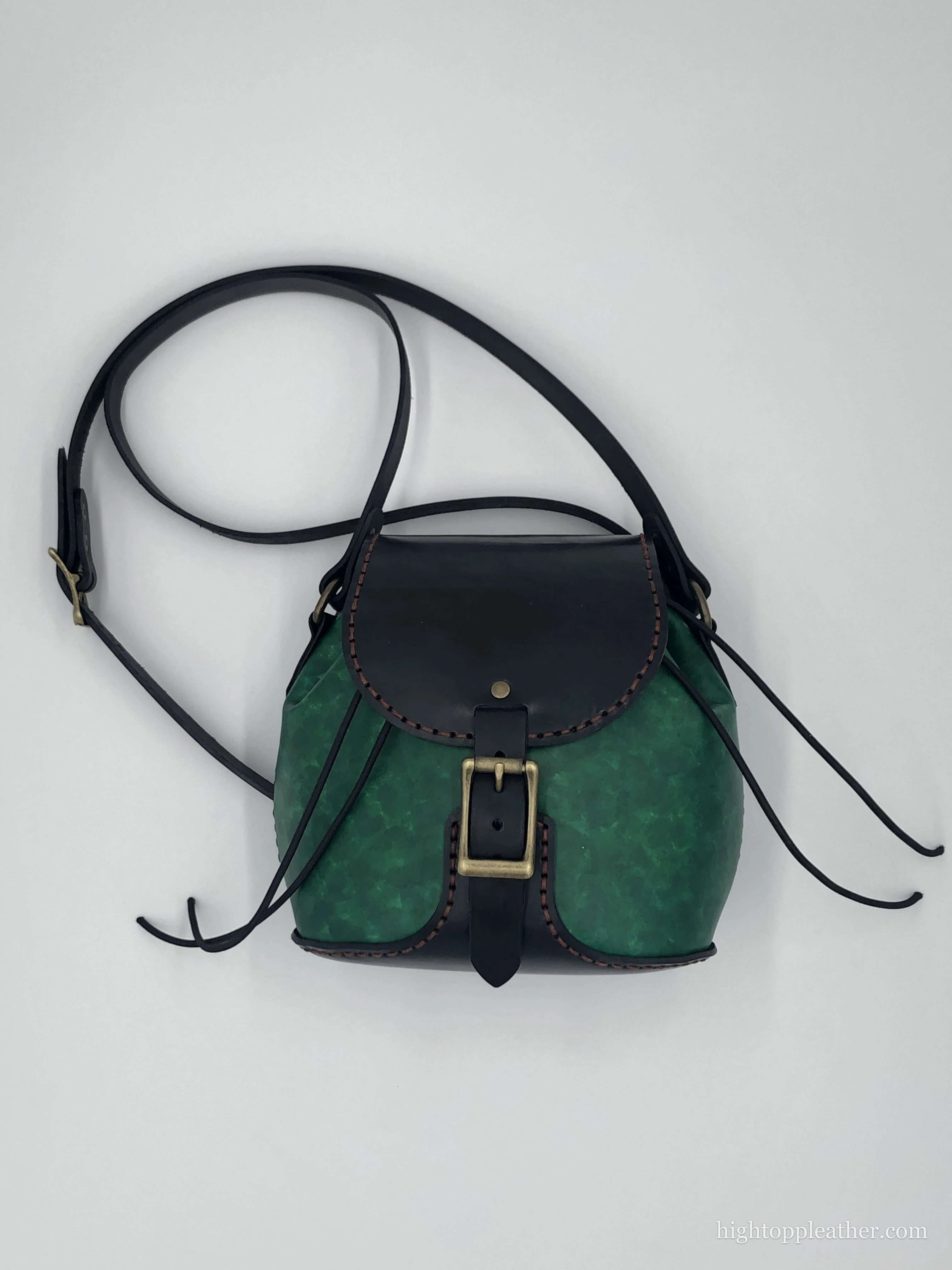 Small crossbody satchel in jade-green dyed leather with a black flap, brass buckle, and saddle stitching.